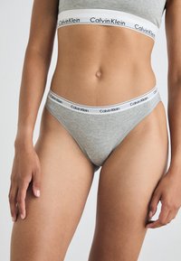 Szürke pamut tanga alsónemű, fehér rugalmas derékpánttal, melyen a "Calvin Klein" márkaneve található. Sima textúra, minimalista varrási részletek.