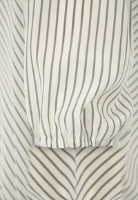 Weiße Stoffbluse mit vertikalen grauen Streifen. Sie hat geraffte Elastikbündchen und eine glatte Textur. Insgesamt entspannte Passform.