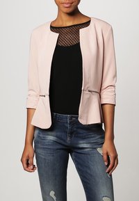 Vaaleanpunainen bleiseri, jossa ei ole kaulusta, kaareva helma ja vetoketjulliset taskut, yhdistetty mustan verkkopaidan ja kuluneiden sinisten farkkujen kanssa.