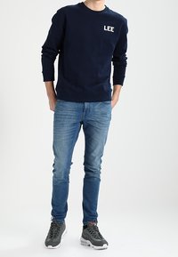Navy blue sweatshirt with "LEE" logo, paired with slim-fit light blue jeans and dark gray athletic shoes. Simple, casual outfit.