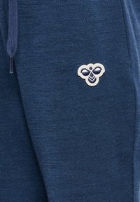 Närbild av marinblå stickade byxor med en vit och marinblå broderad bi-logga nära fickan och en blå dragsko.