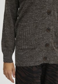 Närbild på en person som bär en brun, lurvig, knäppt kofta med stora fickor och mörka mönstrade byxor.