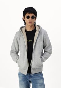 Grå lynlåse-hoodie med en kontrasterende beige fleece-foring, der har en kængurulomme og et logo-emblem på brystet.