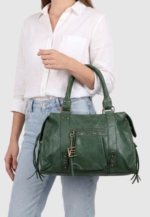 Donna che indossa una camicia bianca e jeans azzurri tiene una borsa a tracolla verde in pelle con tasca frontale e cerniera metallica.