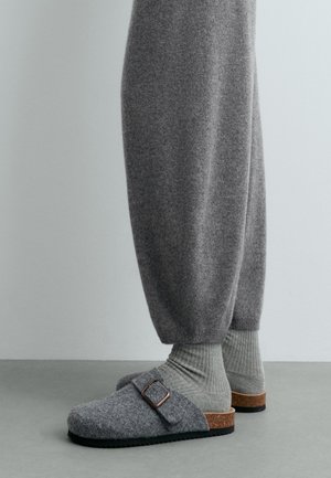 Personne portant un pantalon en laine gris, des chaussettes grises côtelées et des sabots en feutre gris à enfiler avec des boucles et des semelles en liège, debout sur un sol gris.