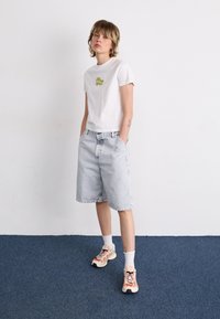 T-shirt blanc à manches courtes avec motif vert, short en denim bleu clair, chaussettes blanches et baskets multicolores. Debout sur un tapis bleu.