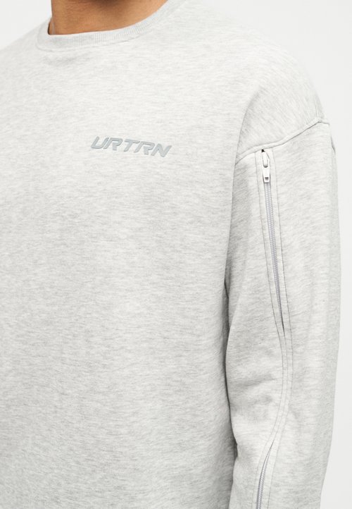 Sweat-shirt gris clair à manches longues, col rond, poches latérales zippées et branding subtil en gris sur la poitrine. Texture douce.