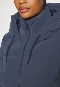 Mørkeblå puffer-jakke med høj krave og snørret hætte. Har en frontlynlås og horisontal quiltet tekstur.
