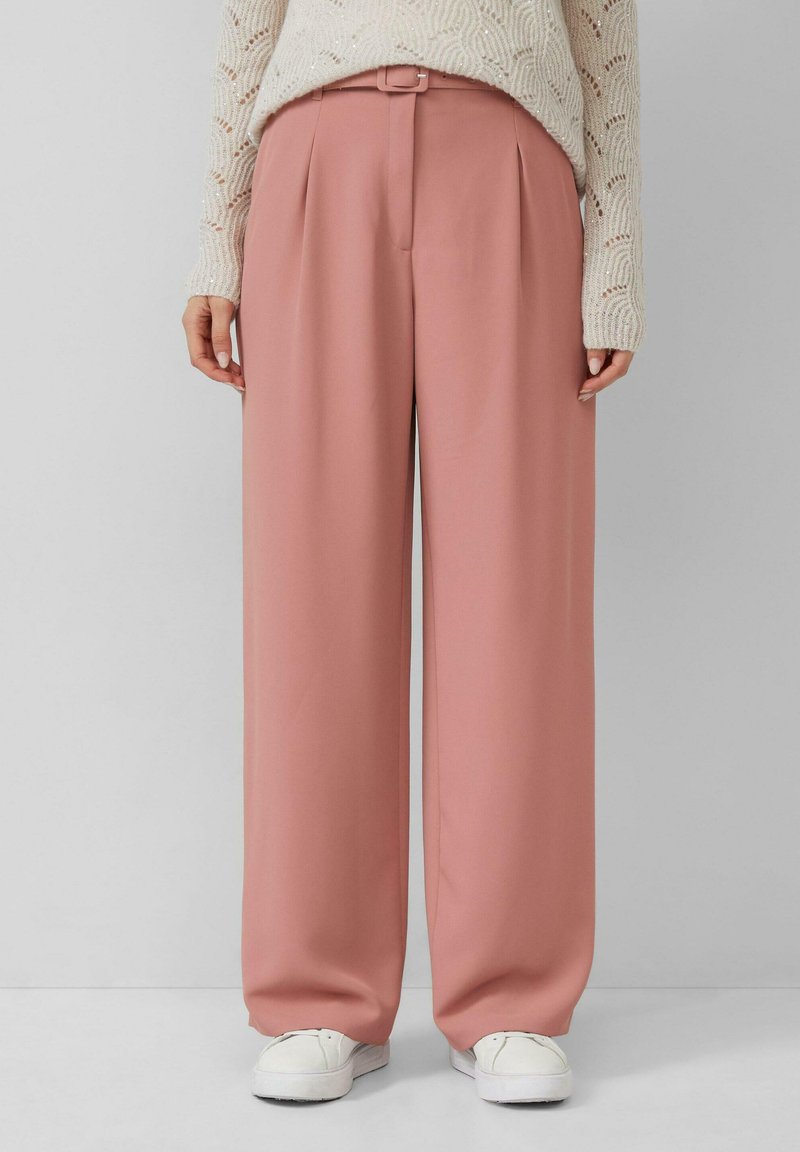 Roze wijde broek met voorplooien, gemaakt van een gladde stof. Gepaarde met een lichtgeweven trui en witte sneakers.
