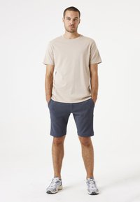 Camiseta de algodón beige de mangas cortas, combinada con pantalones cortos azul marino. El modelo lleva zapatillas ligeras, de pie en un fondo neutro.