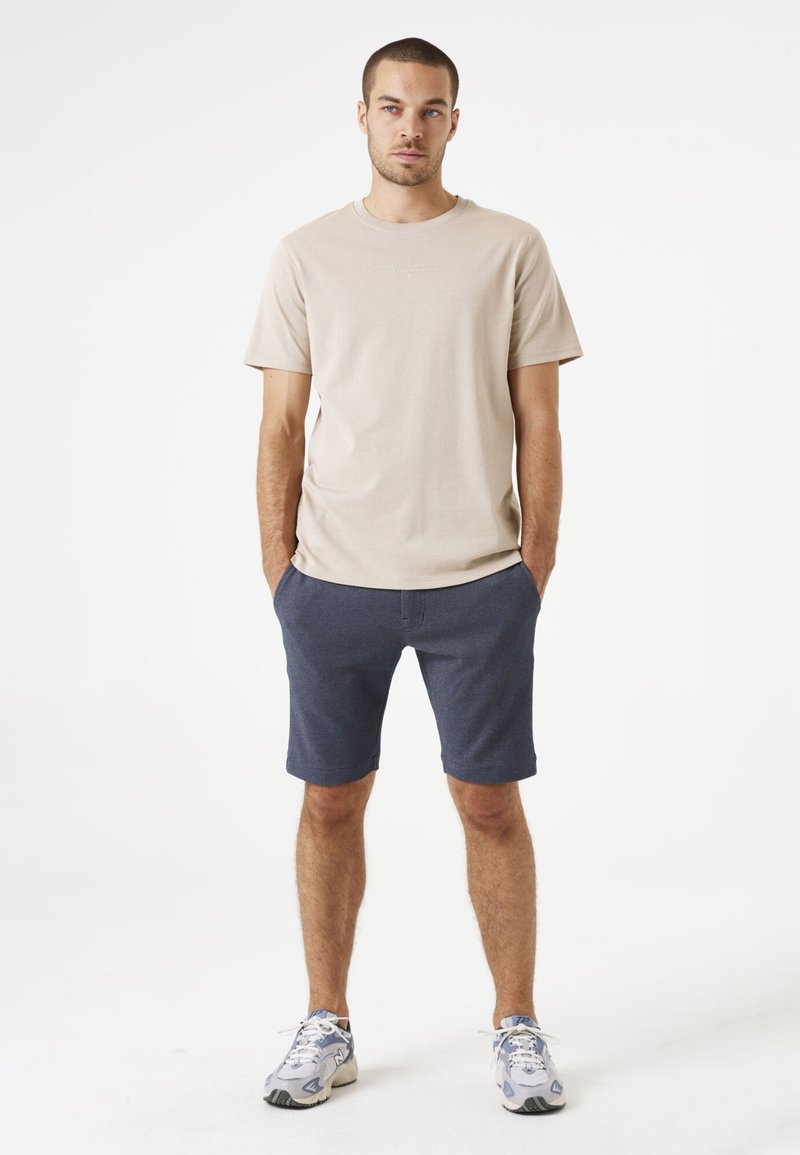 Camiseta de algodón beige de mangas cortas, combinada con pantalones cortos azul marino. El modelo lleva zapatillas ligeras, de pie en un fondo neutro.