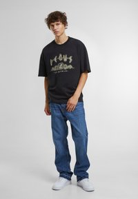 Zwarte oversized t-shirt met grafische print, gecombineerd met losse blauwe denim jeans en witte sneakers. Minimalistisch ontwerp en casual stijl.
