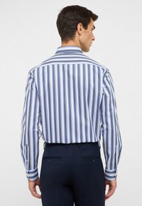 Camicia a righe con righe verticali blu e bianche, con polsini a bottoni e colletto classico, abbinata a pantaloni eleganti blu navy, vista da dietro.