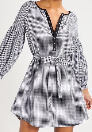 Kobieta ubrana w czarno-białą sukienkę w kratę gingham z bufkowatymi długimi rękawami, przewiązanym paskiem w talii oraz czarną lamówką z guzikami przy dekolcie.