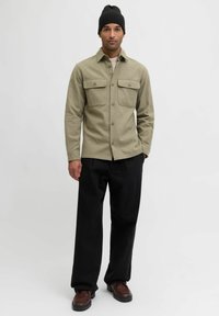 Olivengrønn skjorte med knapper foran og to brystlommer, kombinert med løse svarte bukser og brune sko, komplettert med en svart lue.