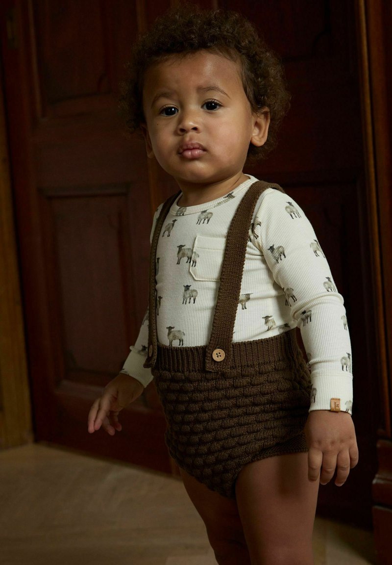 Petit enfant aux cheveux bouclés portant une chemise blanche à manches longues avec des imprimés de chiens et des bretelles tricotées marron, debout à l'intérieur devant des portes en bois.
