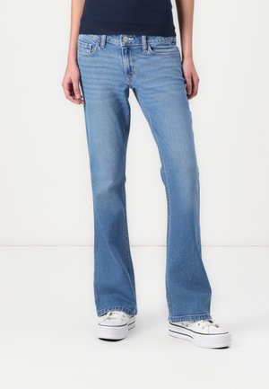 Osoba nosící světlé modré džíny bootcut a bílé tenisky na platformě stojí před bílým pozadím.