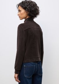 Brun manchesterjacka med hög krage, ribbat mönster och kort design. Matchad med blå denimjeans, sedd bakifrån.