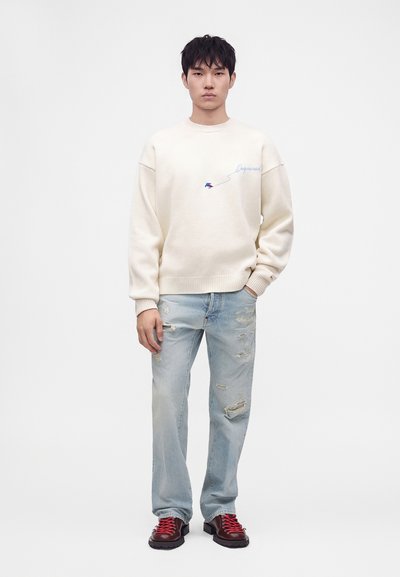 Jeune homme portant un sweat crème, un jean bleu clair déchiré et des bottes rouge foncé avec des lacets rouges, debout devant un fond blanc uni.