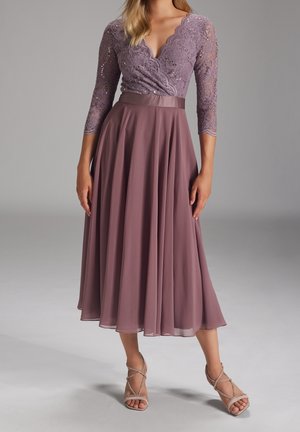 Femme portant une robe en dentelle mauve style portefeuille avec manches trois-quarts et jupe midi fluide, associée à des sandales à talons hauts et à lanières.