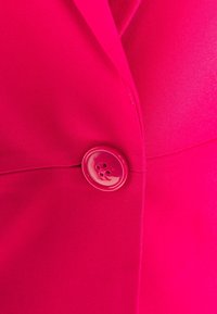 Ljusrosa jacka med en stor matchande knapp fäst fram, som visar en del av kragen.