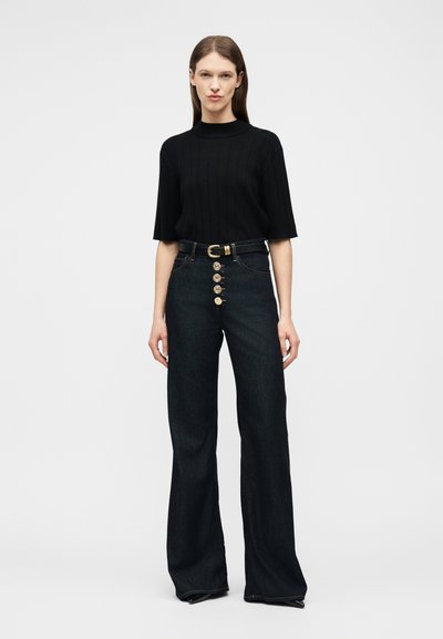 Γυναίκα με ίσια καστανά μαλλιά που φοράει μαύρο ριμπ κοντομάνικο τοπ και μαύρο τζιν ψηλόμεσο με χρυσά κουμπιά και μαύρη ζώνη.