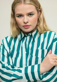 Camisa de botones de algodón con rayas verticales en color teal y blanco, con dos bolsillos de parche y cuello. Textura suave.