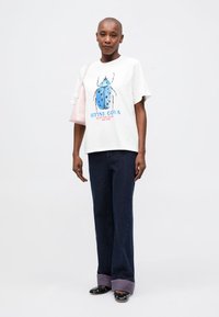 Camiseta blanca con un gráfico de escarabajo en azul y negro, combinada con unos pantalones vaqueros oscuros con puños morados y zapatos negros. Llevando un bolso rosa.