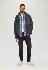 Grijze fleecetrui met capuchon, met een donkergrijze borstzak. Gelaagd over een blauw geruit overhemd. Zwarte spijkerbroek en lichte sneakers maken de outfit compleet.