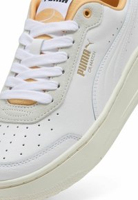 Baskets en cuir blanc avec un accent en suède gris, présentant des perforations, des lacets blancs et un logo doré sur le côté. Col doux et texturé.