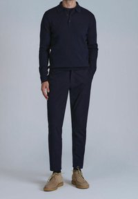 Polo à manches longues côtelé bleu marine associé à un pantalon bleu marine ajusté et des chaussures à lacets en daim beige, présenté sur un fond neutre.