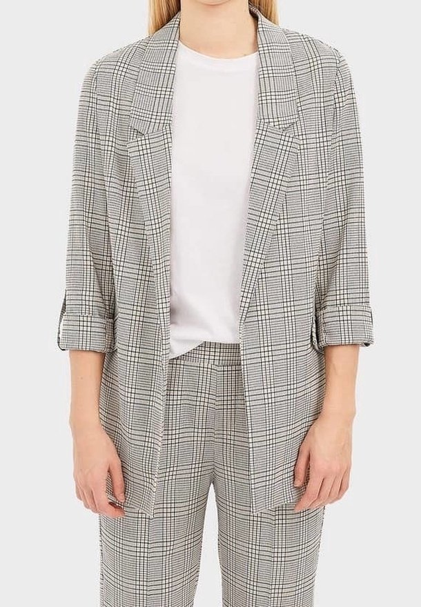Femme portant un blazer à carreaux gris avec les manches retroussées sur une chemise blanche, associé à un pantalon à carreaux assorti.