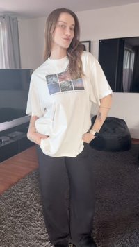 Weißes T-Shirt mit grafischem Druck, kurzen Ärmeln und lockerem Schnitt, kombiniert mit schwarzen, weit geschnittenen Hosen. Im Hintergrund ist ein modernes Wohnzimmer zu sehen.