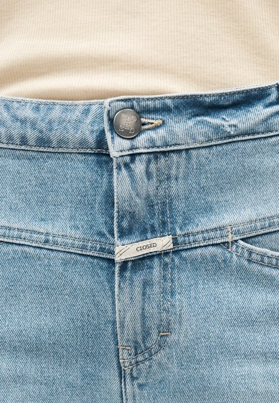 Jean en denim bleu clair avec une fermeture à boutons, des poches avant, et une étiquette cousue portant le mot "CLOSED". Le tissu présente une texture usée.