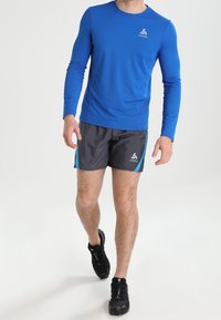 Camisa atlética de manga larga color azul con tejido texturizado, combinada con pantalones cortos grises que tienen acentos en turquesa y un logo. Zapatillas de correr negras visibles.