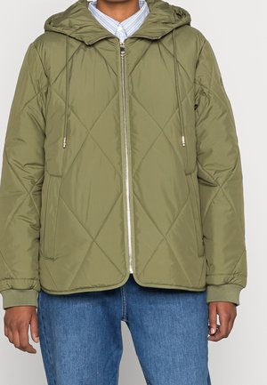 Personne portant une veste matelassée kaki à capuche avec fermeture éclair à l'avant, poches latérales et jean bleu, debout devant un fond uni.