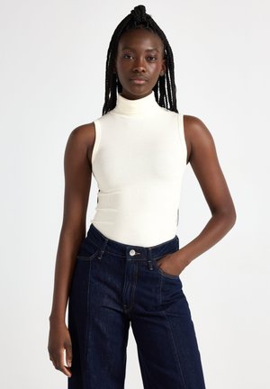 Top in crema a coste senza maniche con colletto alto abbinato a jeans blu scuro a vita alta, dotati di bottone e tasche frontali.