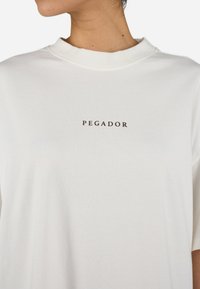 Camiseta de algodón blanca con cuello redondo acanalado, con la palabra "PEGADOR" impresa en texto marrón oscuro en el centro del pecho. Mangas cortas.