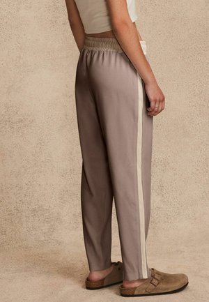 Personne portant un pantalon de survêtement beige avec une bande latérale claire et des sandales marron à enfiler, debout devant un fond beige texturé.