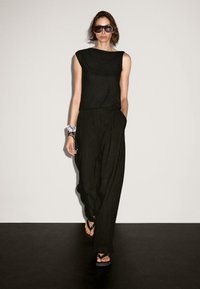 Black sleeveless top and loose-fitting black trousers, paired with flip-flops. Model wears oversized sunglasses. Smooth fabric texture.