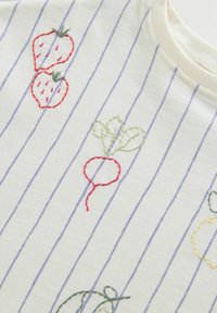 Tissu blanc à rayures verticales bleues avec des fraises rouges brodées, un radis rouge avec des feuilles vertes et un contour partiel de légume jaune.
