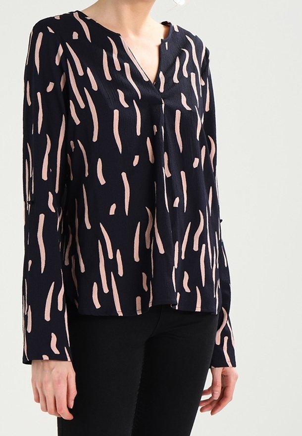 Femme portant un chemisier noir à manches longues avec des stries abstraites roses et un pantalon noir sur un fond clair uni.