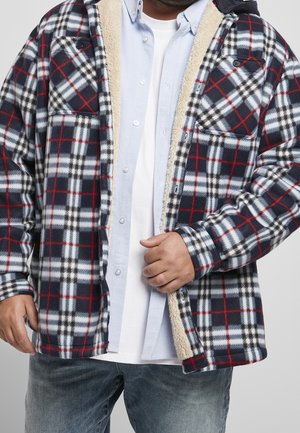 Mann trägt eine blau-weiß-rot karierte Fleecejacke mit beigefarbenem Innenfutter über einem hellblauen Hemd mit Knöpfen und einem weißen T-Shirt, kombiniert mit blauen Jeans.