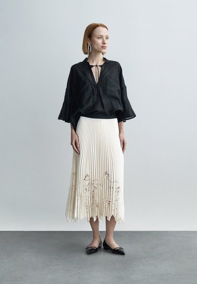 Μαύρο λινό τοπ με φαρδιά μανίκια, συνδυασμένο με πλισέ εκρού φούστα με κεντήματα λουλουδιών και μαύρες μυτερές μπαλαρίνες.