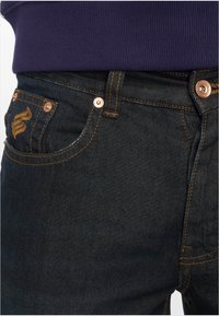 Mørk denim jeans med gyldne sømmer, kobbernitter, en myntlomme og en subtil merkevarelogo på baklommen.