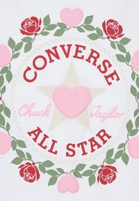 Bielej grafike s červeným textom "CONVERSE ALL STAR" obklopeným zelenými listami, ružovými srdcami a červenými ružami, umiestneným na béžovej hviezde.