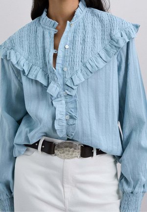 Blouse bleu clair à manches longues en tissu texturé avec des détails volantés, rentrée dans un pantalon blanc avec une ceinture foncée et une grande boucle ovale.