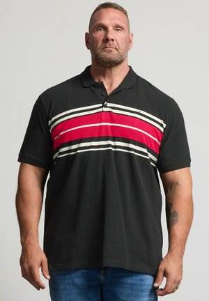 Mand med kort hår og skæg iført en sort polo-shirt med røde og hvide horisontale striber og blå jeans, stående mod en lys baggrund.