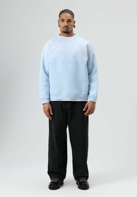 Sweatshirt bleu clair avec petits points noirs, associé à un pantalon noir ample et des chaussures noires. Texture lisse et coupe décontractée.
