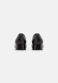 Zapatos de cuero negro con un diseño redondeado, tacones bajos en bloque, textura suave y un detalle de costura mínima, vistos desde la parte trasera.